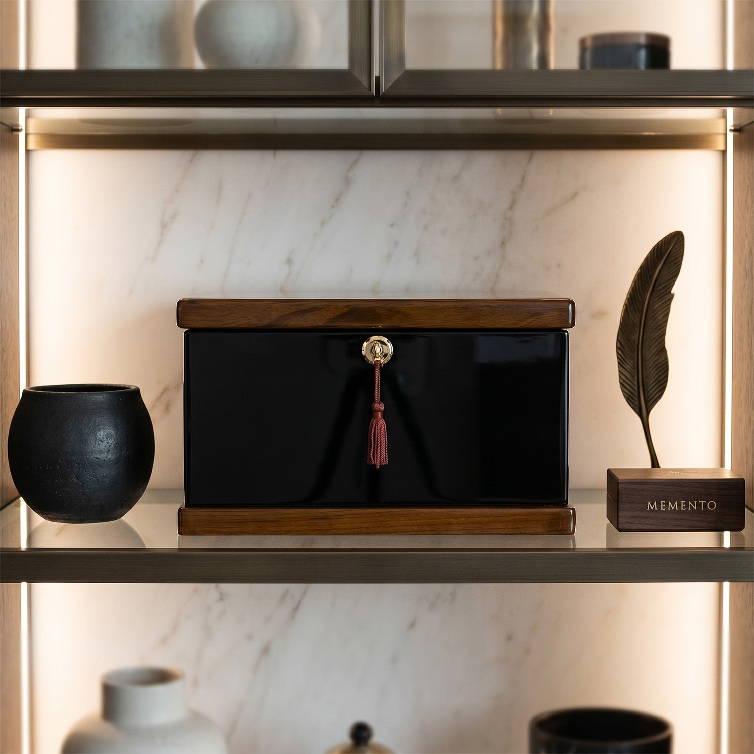 Black rectangular urn box with wooden top and tassel on a shelf with decorative items.